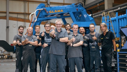A group of men are standing together with a blue excavator behind them and they all have thumbs up signs.