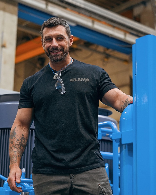 Man wearing a shirt with 'GLAMA' logo standing next to blue equipment.