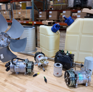The image shows a collection of various mechanical components and parts laid out on a table with white background shelves filled with boxes in the.