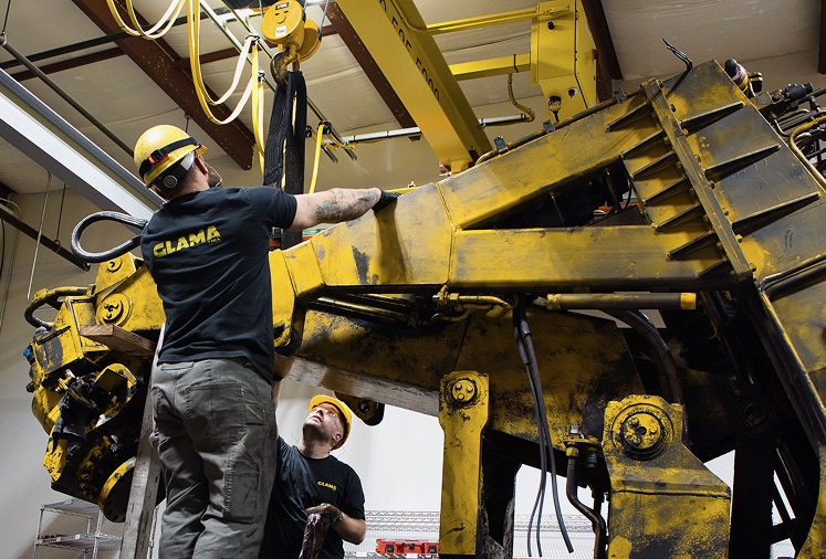 Two workers wearing yellow hard hats and safety vests work with a large mechanical machine in an industrial setting.